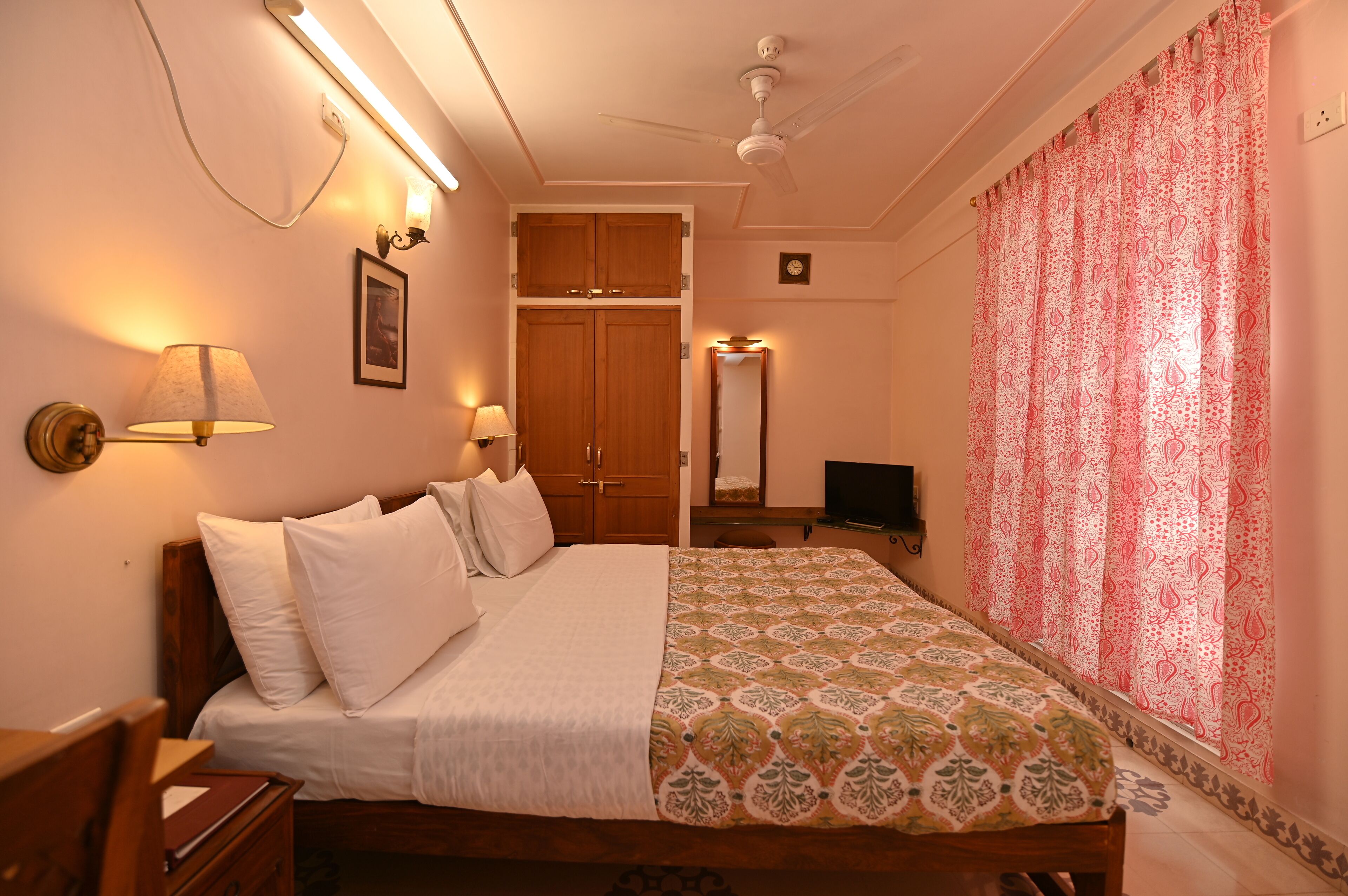 A neatly arranged bedroom with a bed, bedside lamps, a wooden wardrobe, a television, and a patterned curtain.