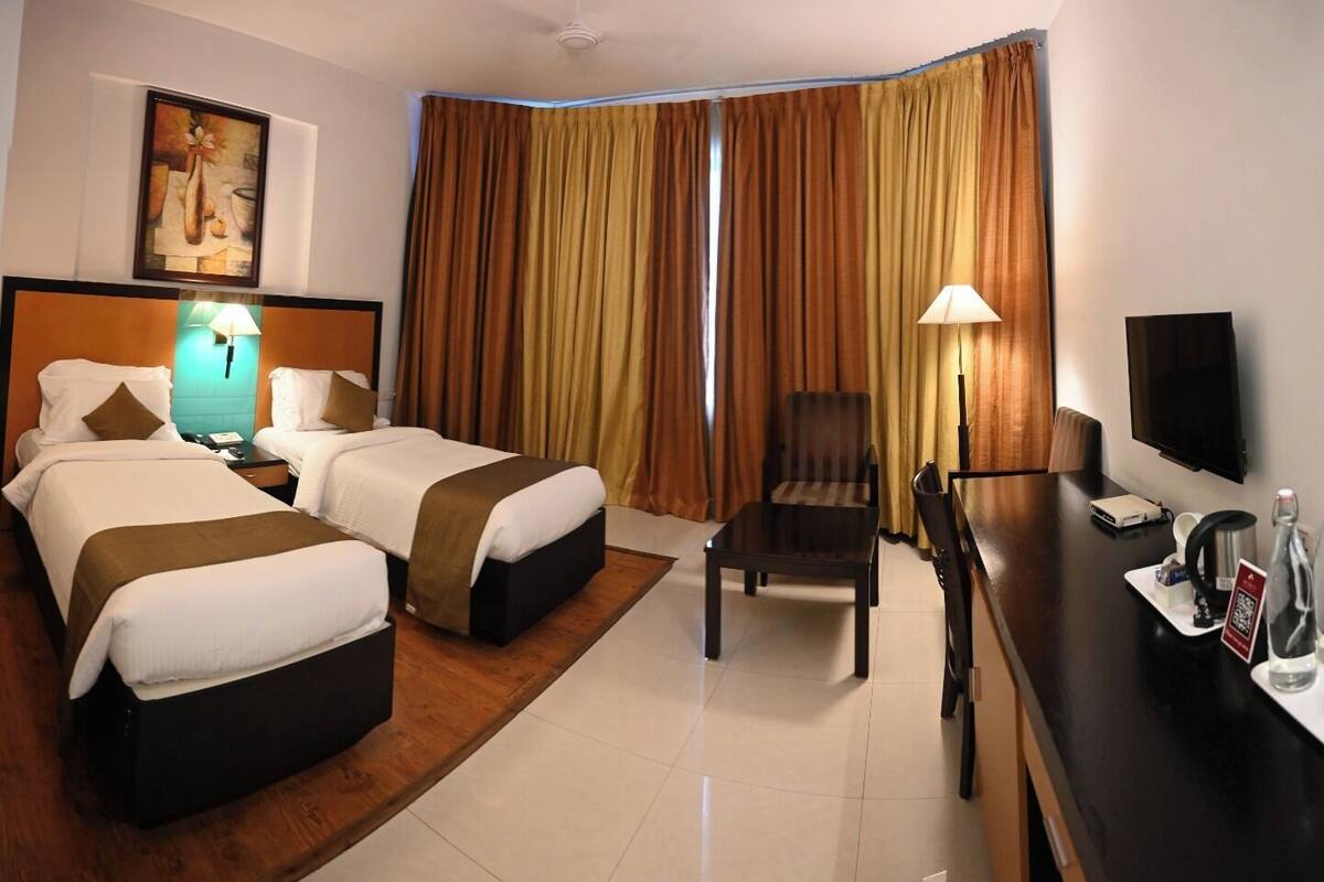 A hotel room with two beds, a desk with a chair, a TV, and a large window with curtains.