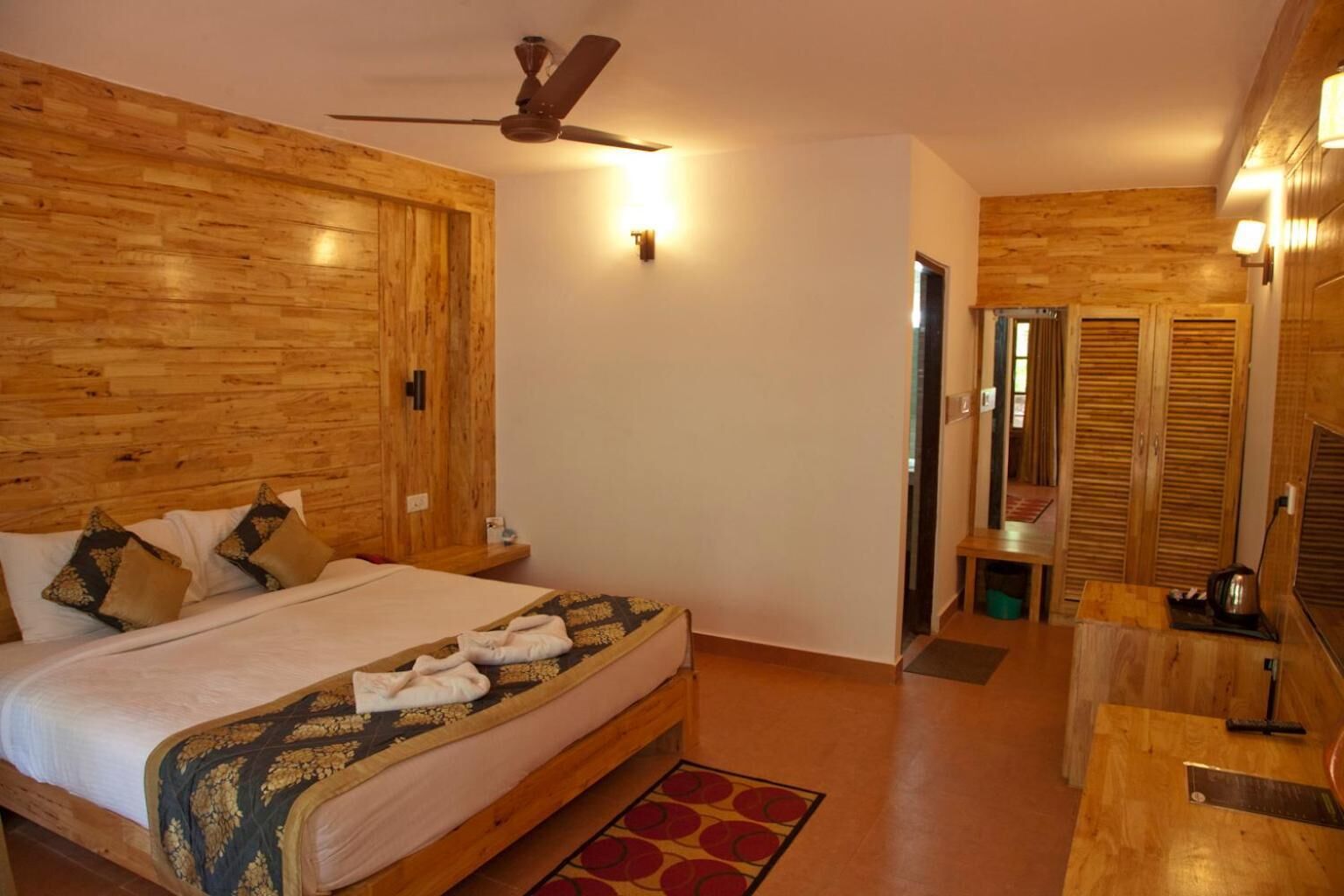 Ein Hotelzimmer mit einem Holzkopfteil, einem Bett mit einem gemusterten Bettbezug, einem Deckenventilator, einem kleinen Tisch mit einem Wasserkocher und einem Holzkleiderschrank.
