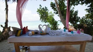 A wooden bed with white linens and a heart-shaped towel arrangement, surrounded by beachside trees and a view of the ocean. - Symphony Palms Beach Resort (Port Blair)