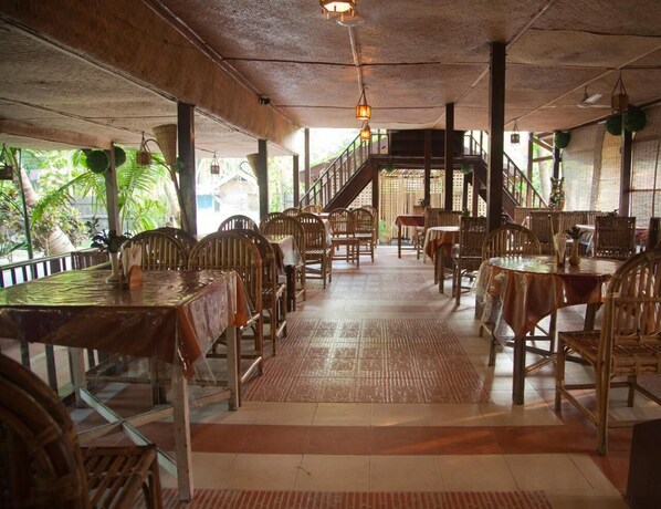 A restaurant with wooden tables and chairs, a staircase, and a view of greenery outside. - Symphony Palms Beach Resort (Port Blair)