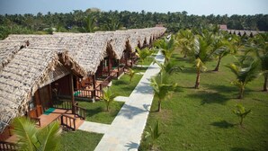 A row of thatched-roof bungalows with outdoor seating, a paved walkway, and palm trees. - Symphony Palms Beach Resort (Port Blair)