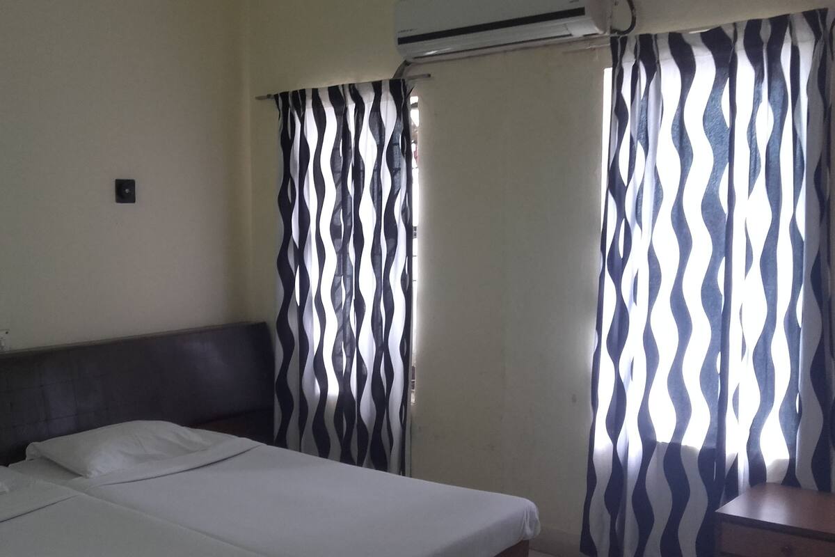 A bedroom with a bed, a nightstand, a wall-mounted air conditioner, and a window with patterned curtains.