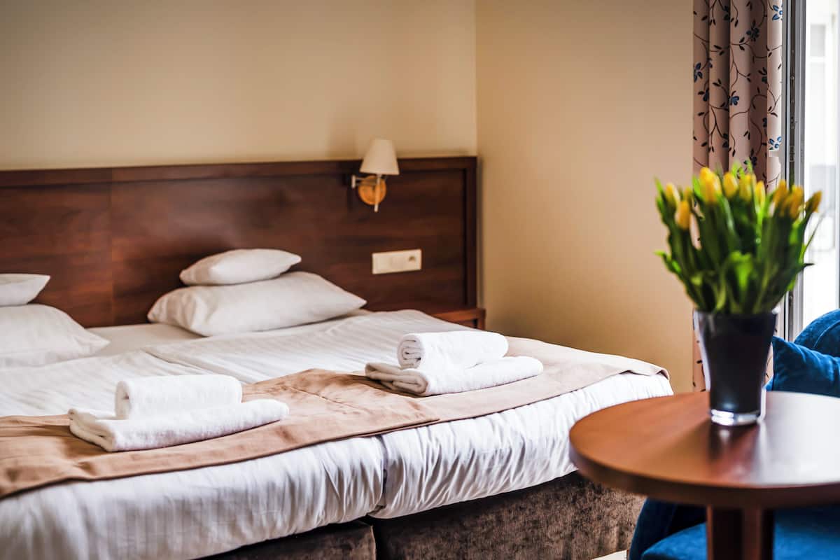 A neatly made bed with white linens and a brown throw blanket, a bedside lamp, and a vase with yellow flowers on a wooden table.