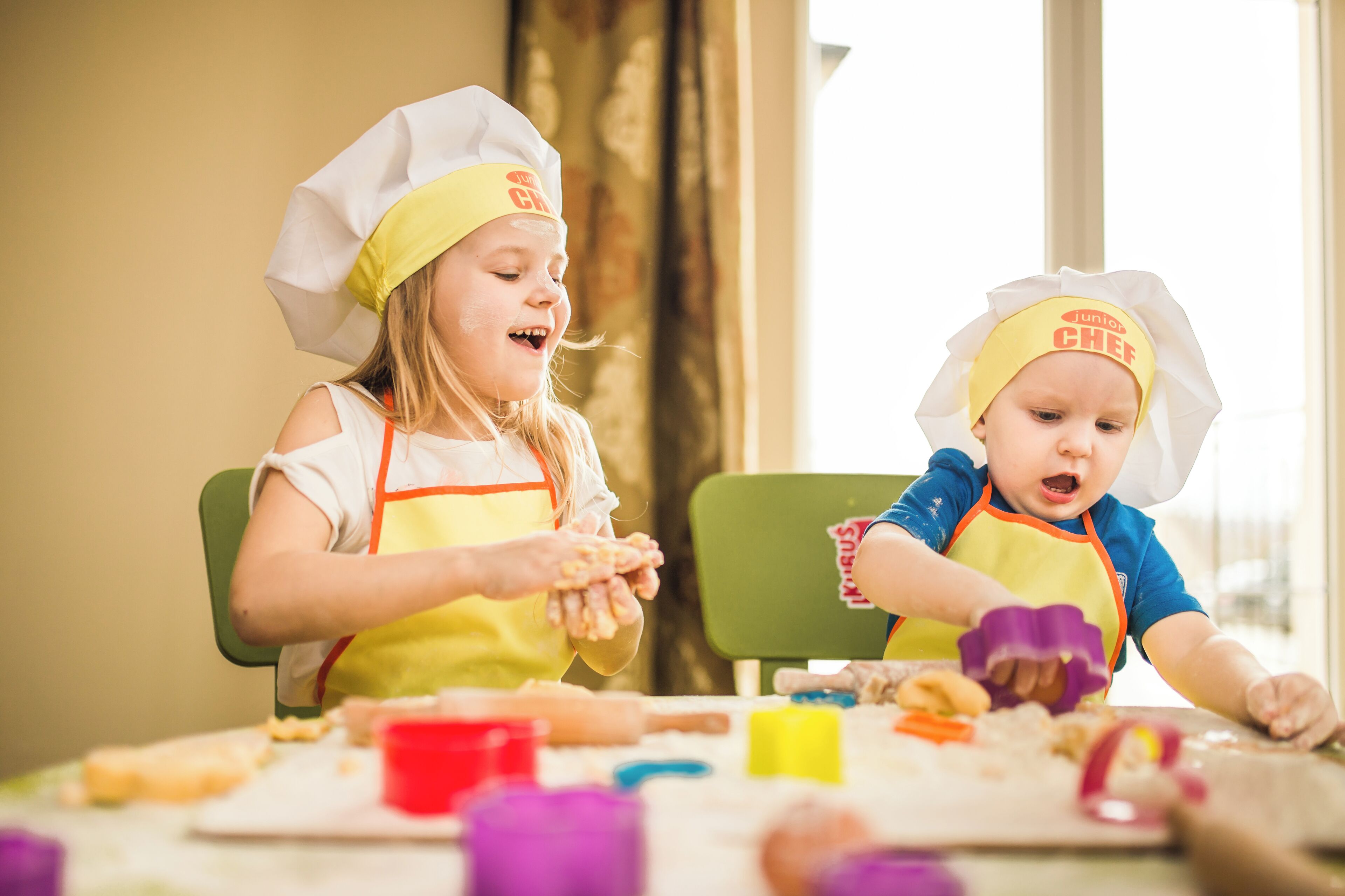 Two children wearing chef hats and aprons, sitting at a table with various baking ingredients and utensils.