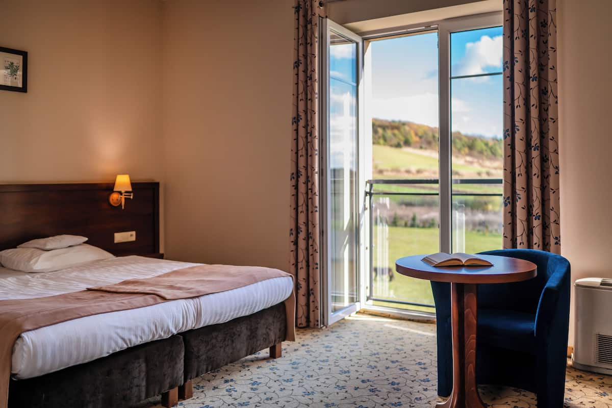 A hotel room with a bed, a bedside table with a book, a chair, and a view of the outdoors through a sliding glass door.