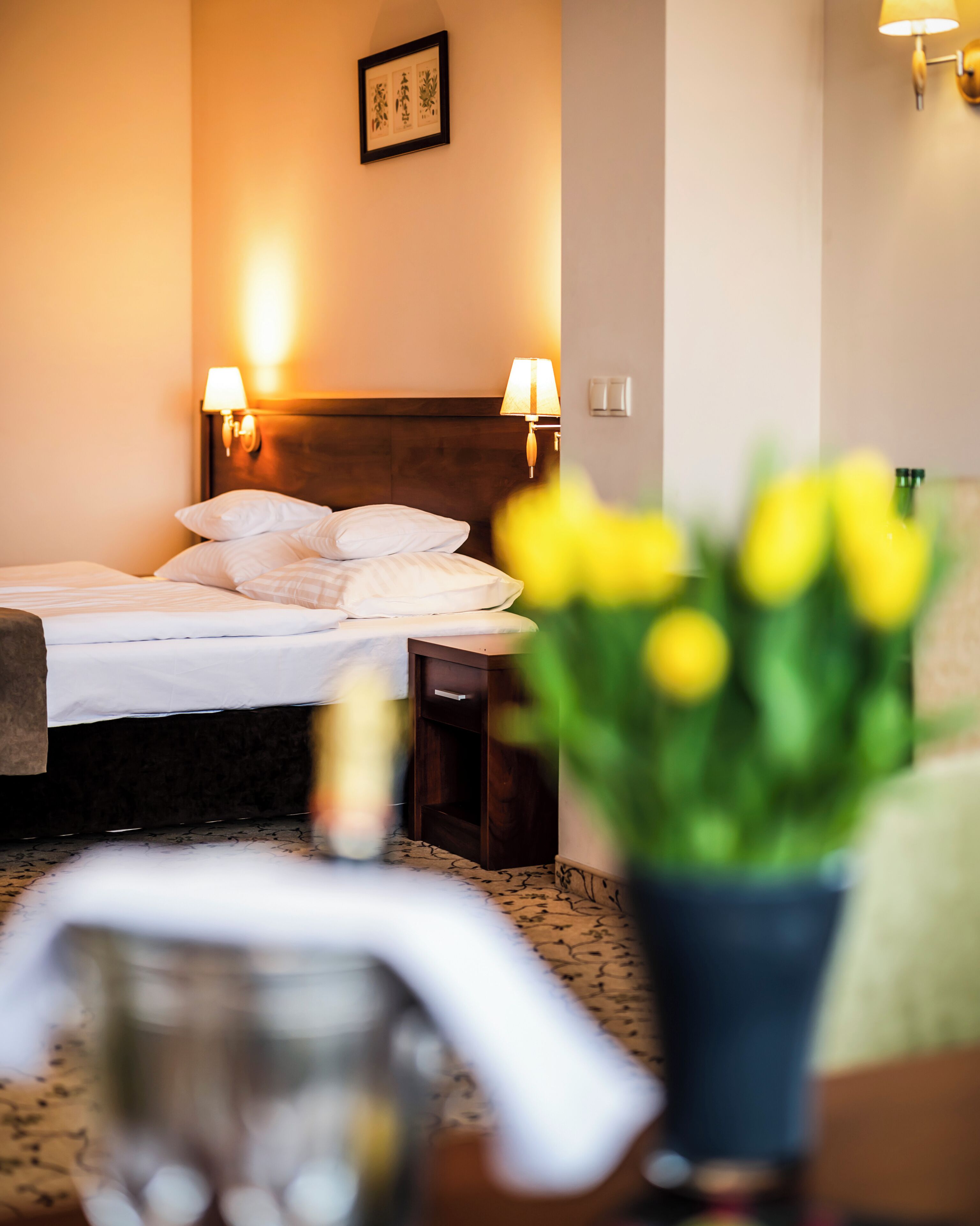Una habitación de hotel con una cama, lámparas de noche, un cuadro en la pared y un florero con flores amarillas.