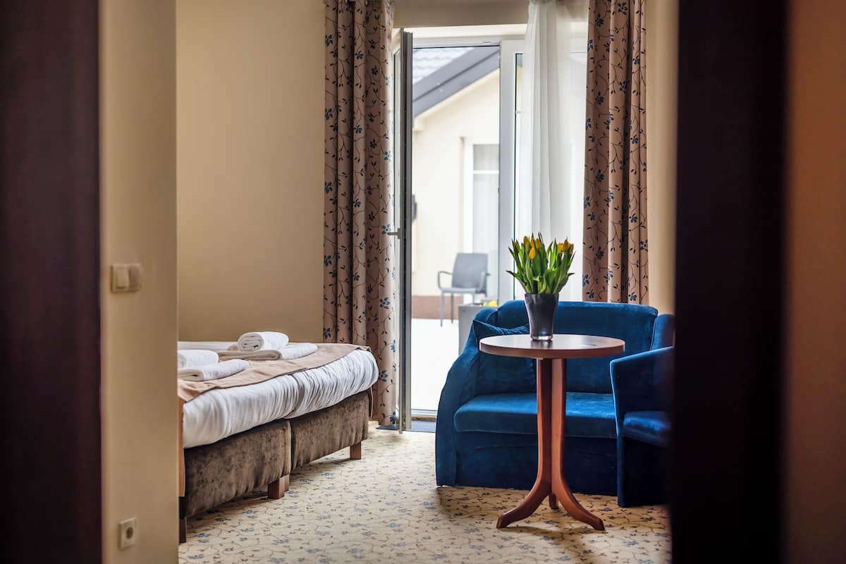 A hotel room with a bed, a blue sofa, a small table with a vase of flowers, and a view of the outside.