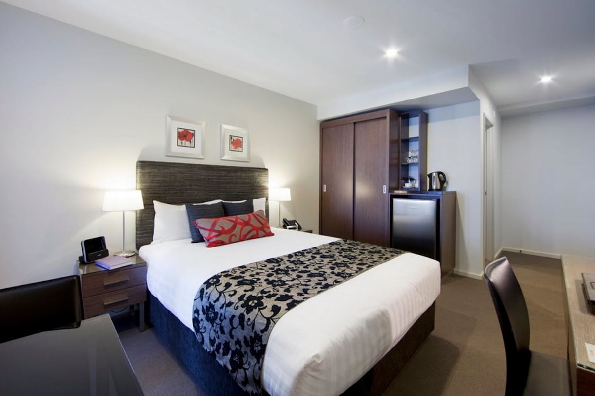 Una habitación de hotel con cama, mesitas de noche, escritorio, silla, armario y kitchenette.