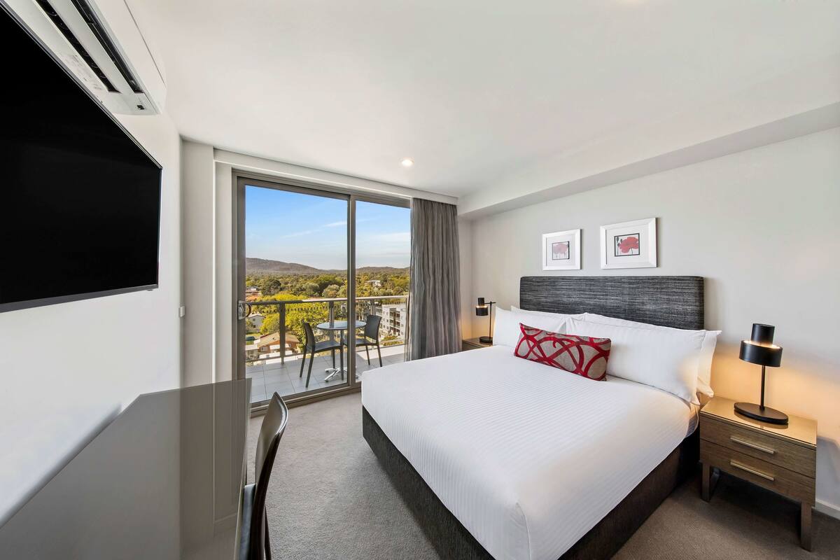 Ein Hotelzimmer mit einem großen Bett, einem Schreibtisch, einem Sessel, einem Fernseher und einem Balkon mit Blick auf die Stadt.