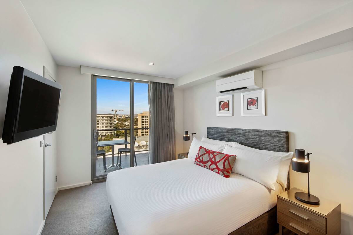 Ein Hotelzimmer mit einem großen Bett, einem an der Wand befestigten Fernseher, einem Balkon mit Tisch und Stühlen und Blick auf die Stadt.