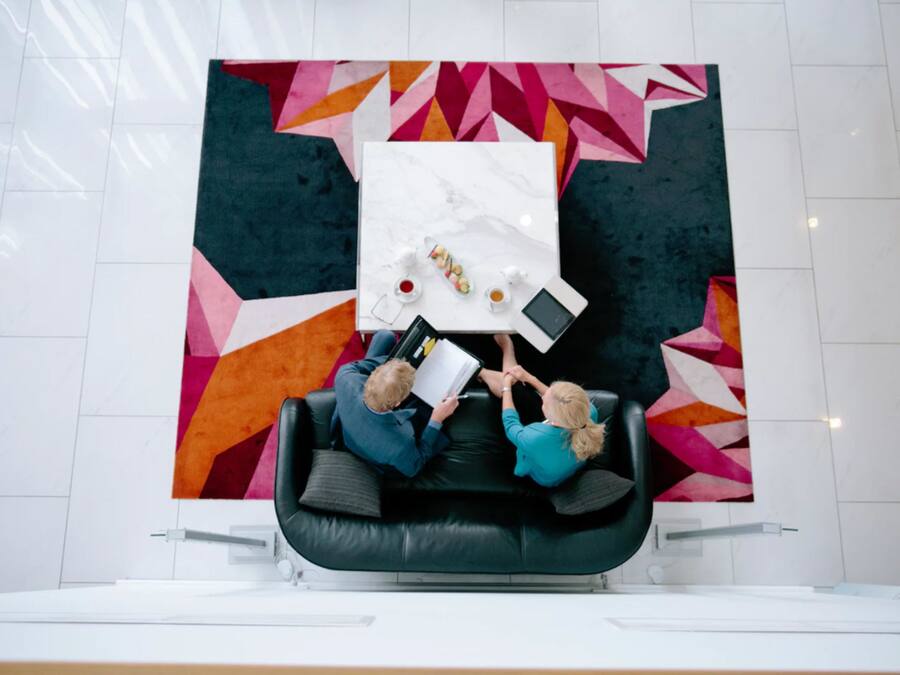 Ein moderner Büroraum mit einer schwarzen Ledercouch, einem weißen Tisch mit einem Tablet und einer Tasse sowie einem farbenfrohen geometrischen Wandmuster.