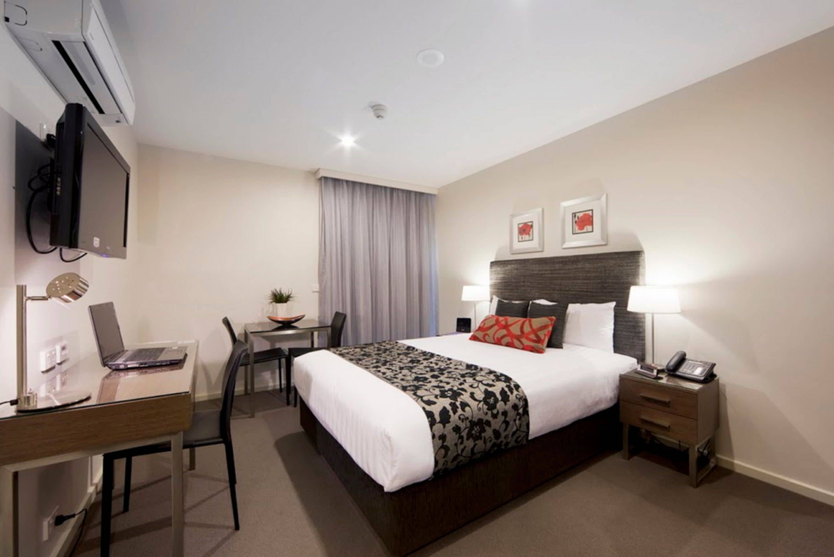 Hotelkamer met een bed, nachtkastje, lamp, bureau met laptop, stoel en een aan de muur bevestigde televisie.