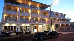 Un hotel de varios pisos con terraza al aire libre y un coche antiguo estacionado al frente.