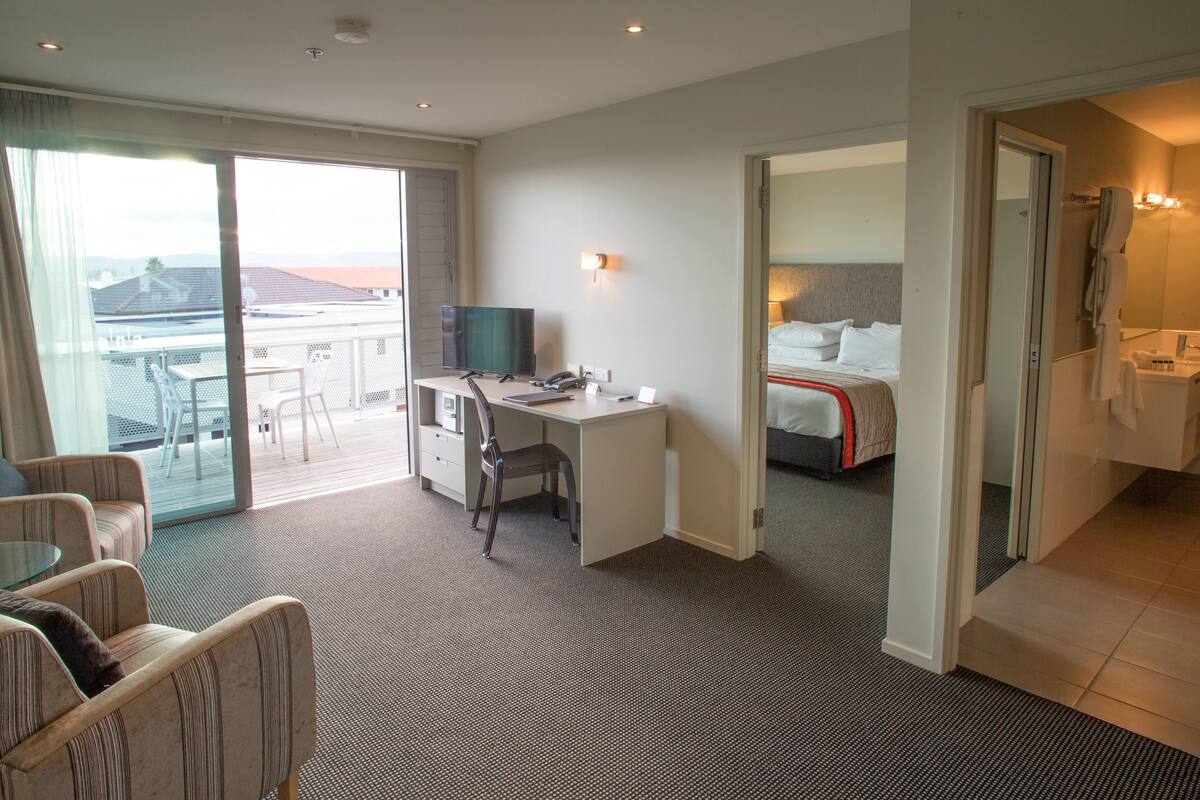 A hotel room with a desk, chair, bed, and a balcony with a view.