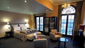 Habitación de hotel con una cama grande, dos sillones, una mesita, un escritorio y vistas a la ciudad a través de amplios ventanales.