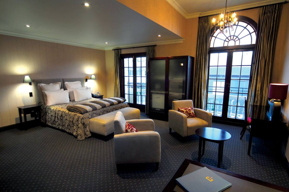 A hotel room with a large bed, two armchairs, a small table, a desk, and a view of the city through large windows.