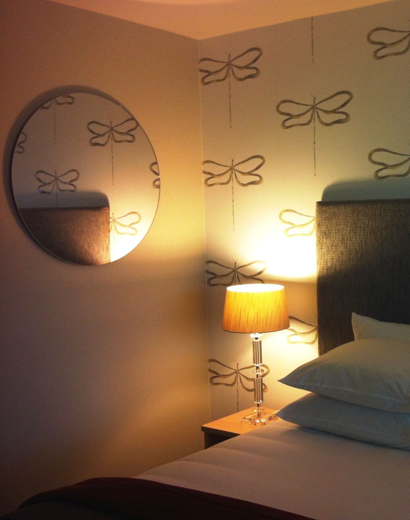 Una habitación de hotel con cama, mesita de noche, lámpara, espejo y papel tapiz con estampado de mariposas.