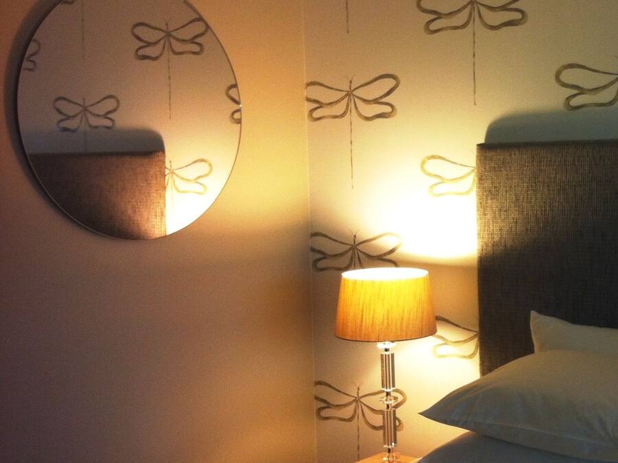 Una habitación de hotel con cama, mesita de noche, lámpara, espejo y papel tapiz con estampado de mariposas.