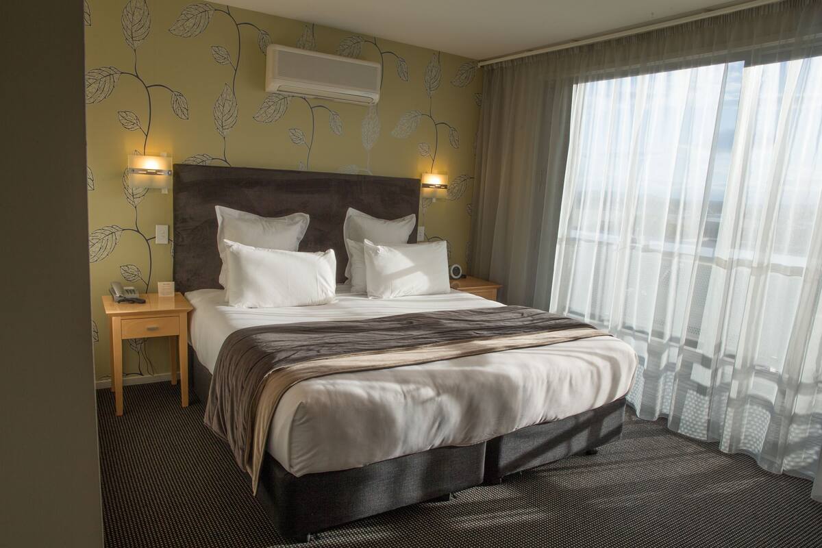 A hotel room with a large bed, two bedside tables, a wall-mounted air conditioner, and a window with sheer curtains.
