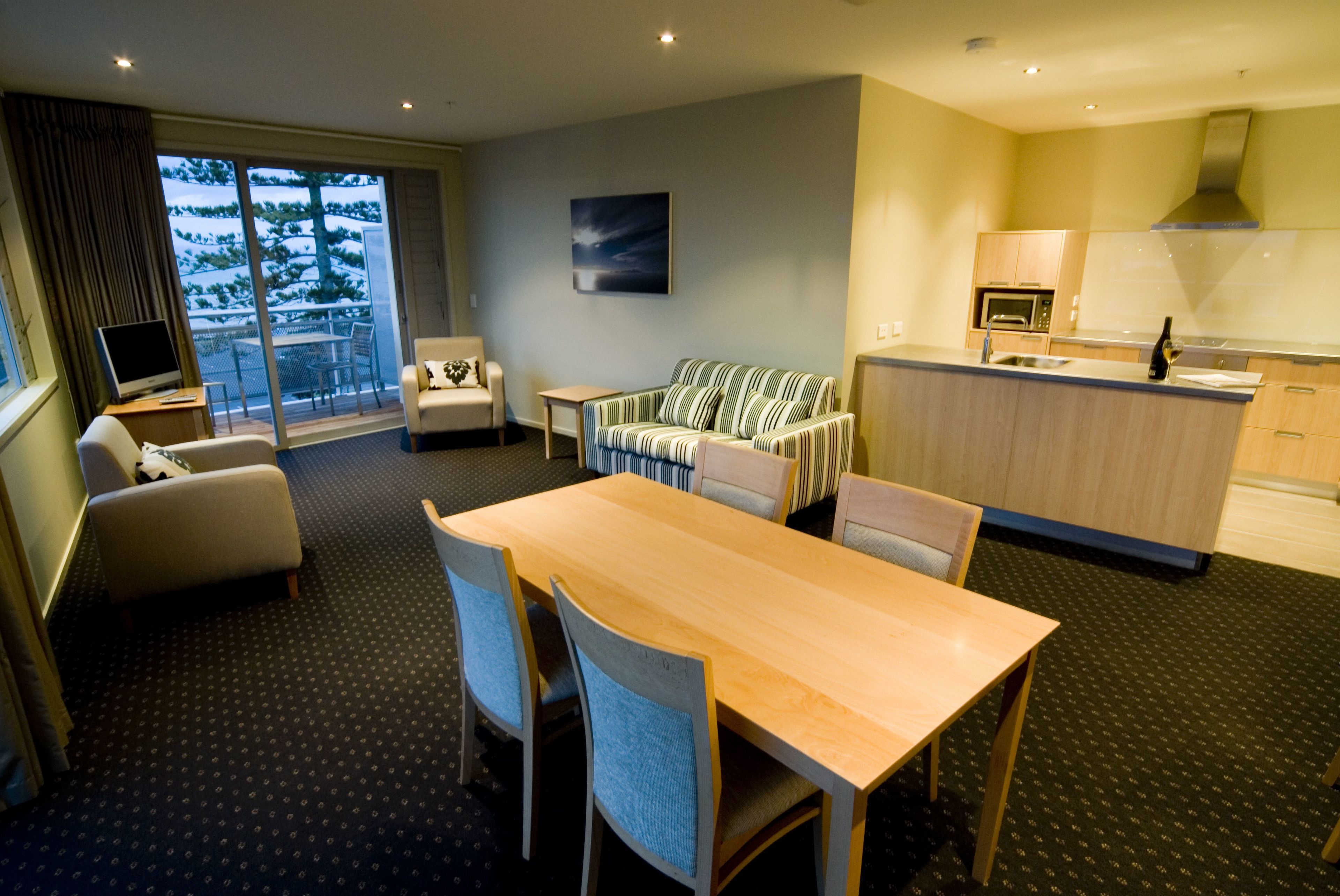 Quarto de hotel com área de refeições, sofá, TV e varanda com vista para árvores.