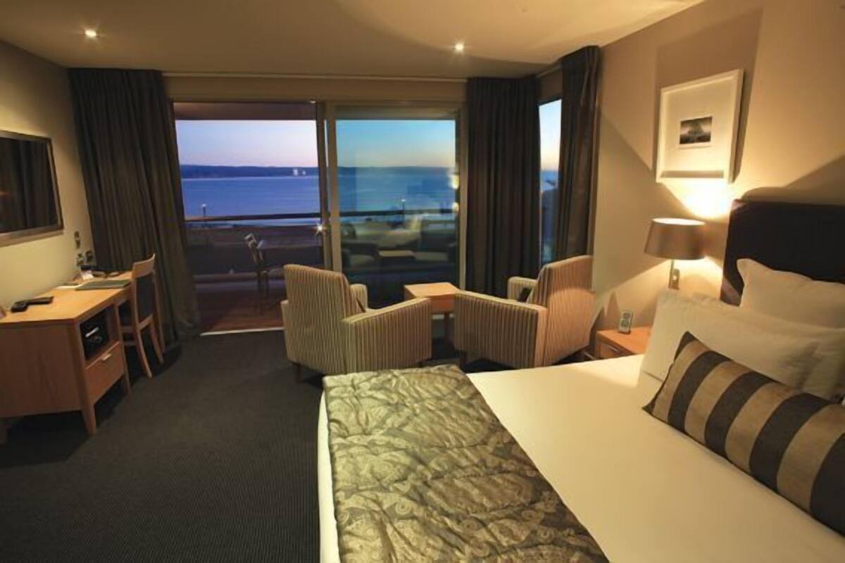 A hotel room with a bed, a desk, a chair, a sofa, and a view of the sea.