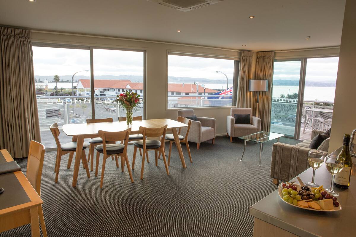 A hotel room with a dining area, a sofa, a table with a wine glass and a plate of grapes, and large windows with a view of the city and mountains.