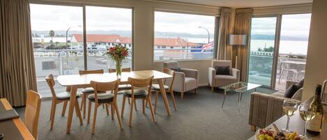 Habitación de hotel con comedor, sofá, mesa con copa de vino y una fuente de uvas, y ventanas amplias con vistas a la ciudad y las montañas.