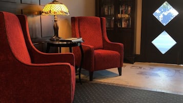 A room with two red upholstered chairs, a small table with a lamp, and a bookshelf.