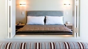 Una cama bien hecha con un cabecero gris, dos almohadas blancas y una almohada a rayas al pie. Hay dos mesitas de noche, una de ellas con una lámpara y un pequeño reloj.