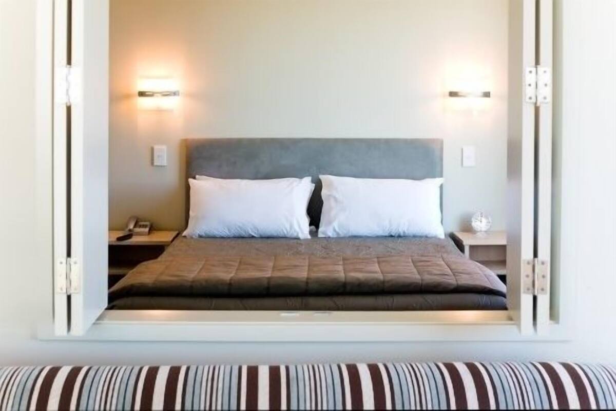 A neatly made bed with a grey headboard, two white pillows, and a striped pillow at the foot of the bed. There are two bedside tables with lamps and a small clock on one of them.