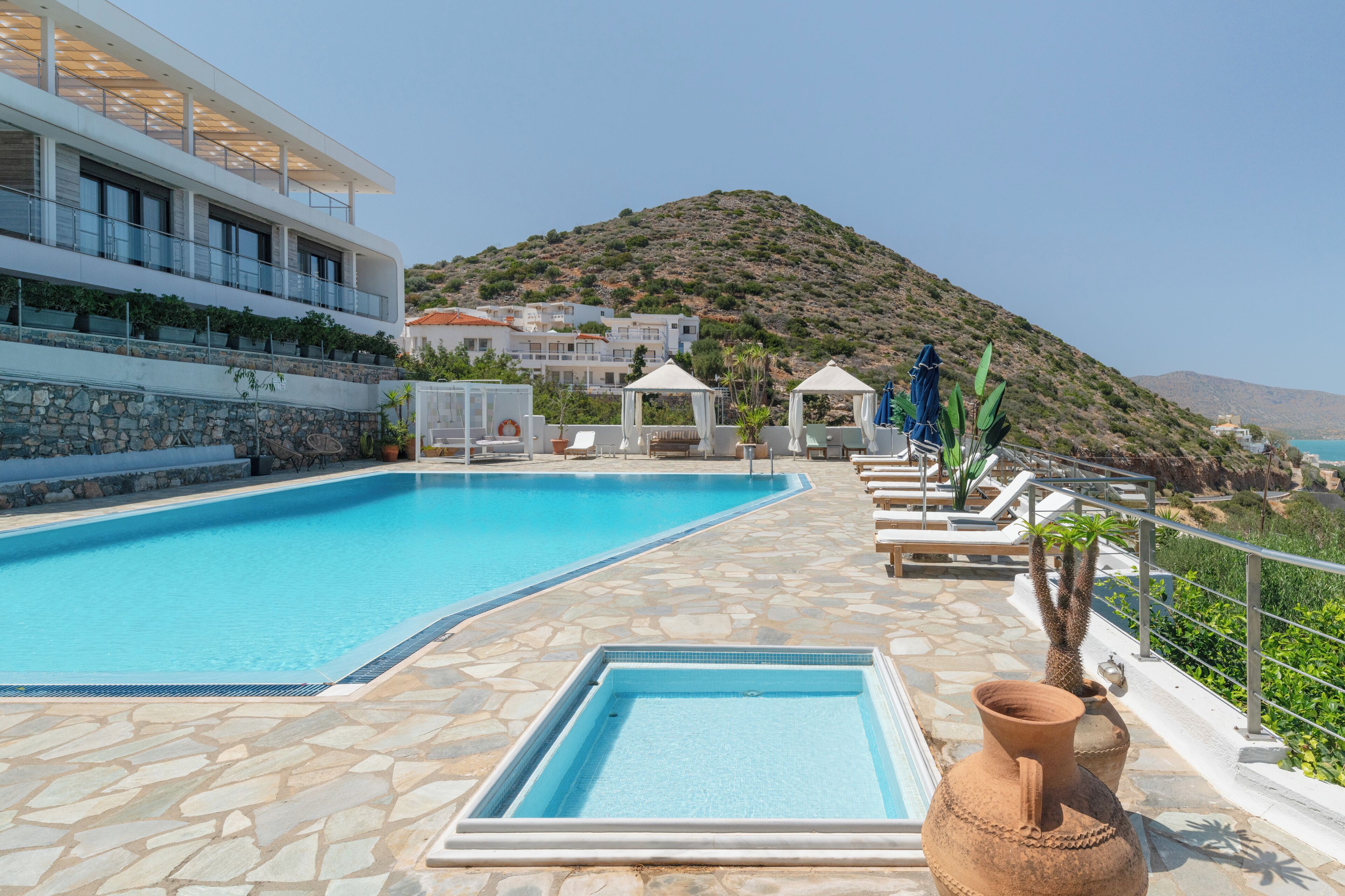 Una piscina con pavimento in pietra, poltrone relax e vista su una collina e edifici sullo sfondo.