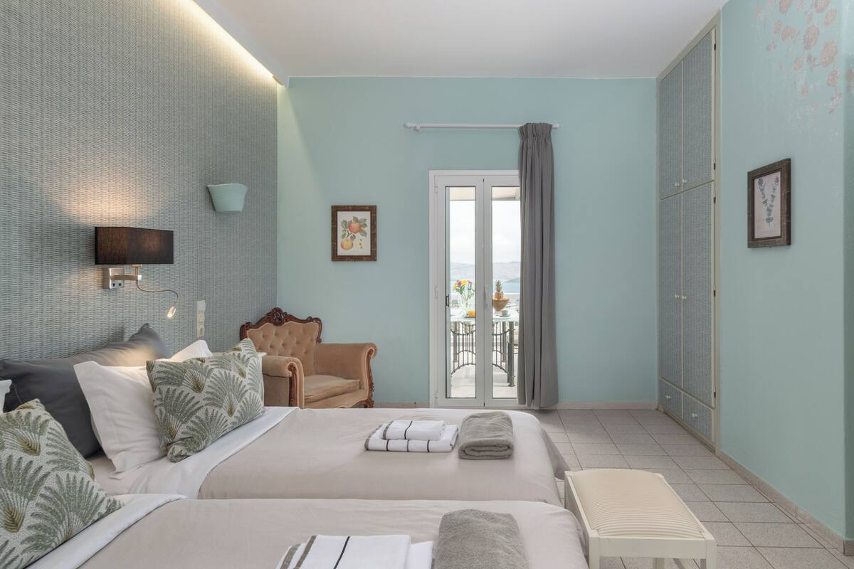 A hotel room with two beds, a sitting area with a chair, a balcony with a table, and a wardrobe.
