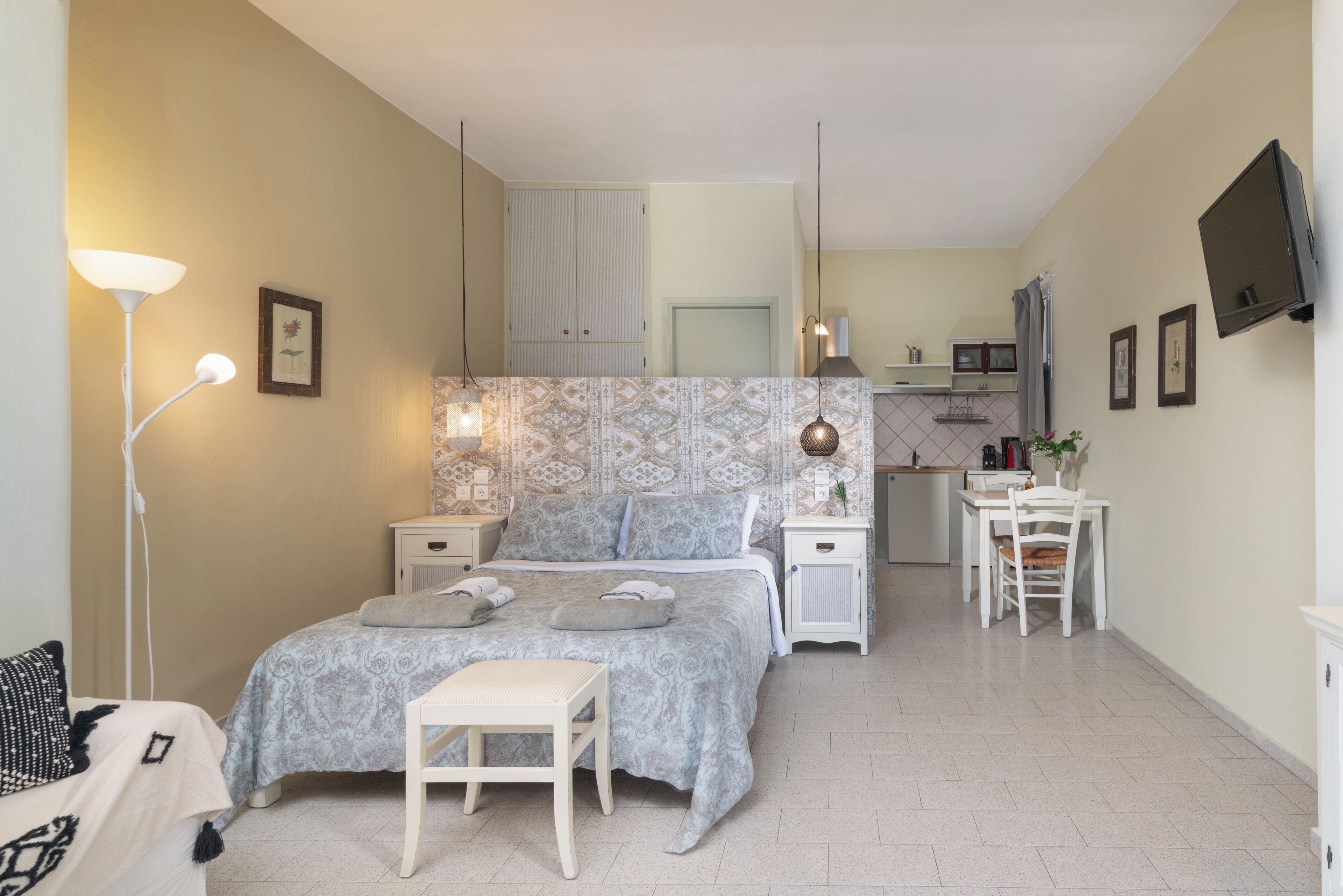 Ein Schlafzimmer mit einem Bett, Nachttischen, einem Schreibtisch und einem Stuhl. An der Wand ist ein Fernseher angebracht und es gibt ein Fenster mit Vorhängen.