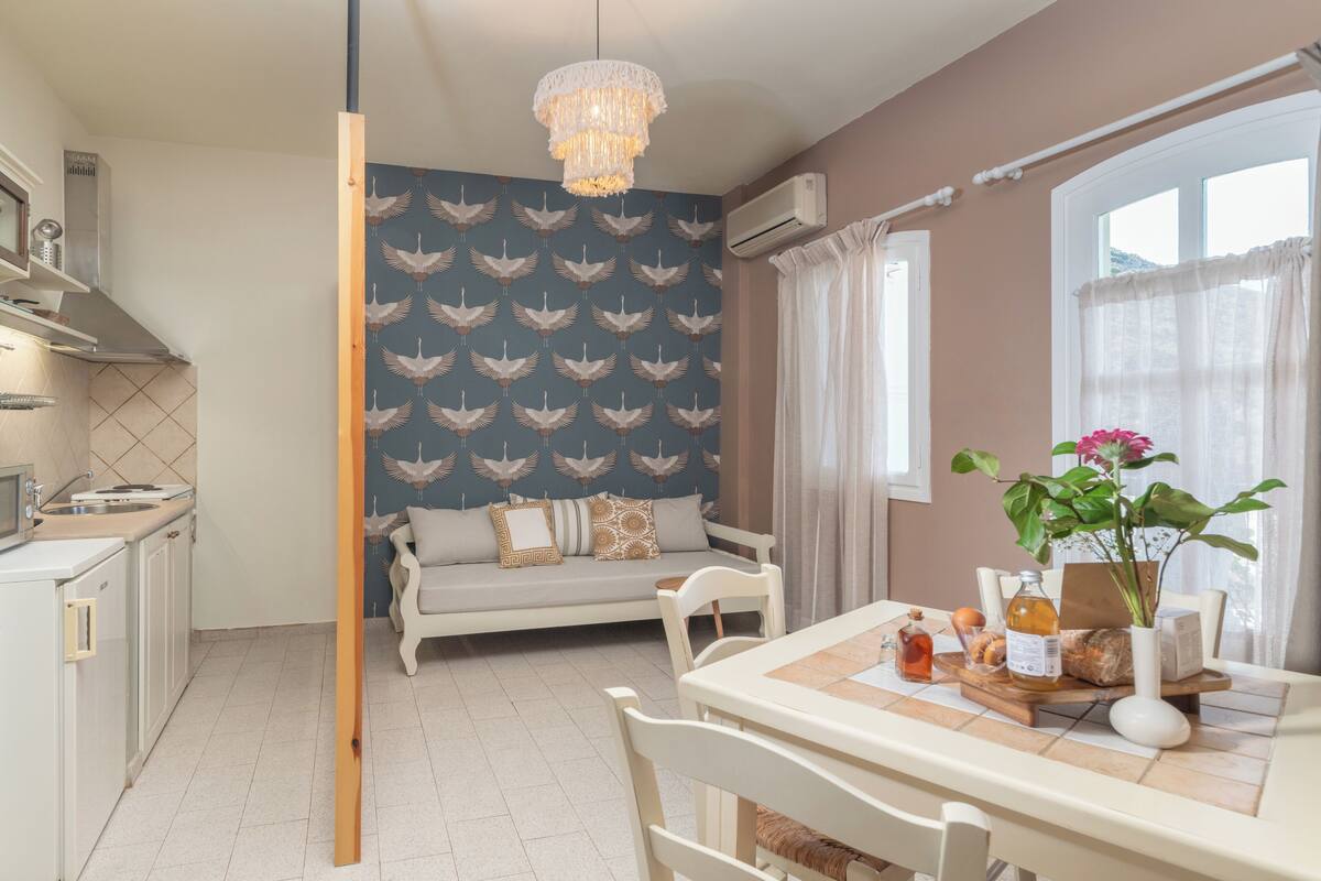 A modern kitchen with white cabinets, a dining table with chairs, a chandelier, and a sofa with patterned wallpaper.