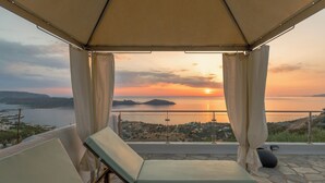 A balcony with a view of the sunset over the sea, a lounge chair, and a stone floor. - Adrakos Apartments - Adults Only (Agios Nikolaos)