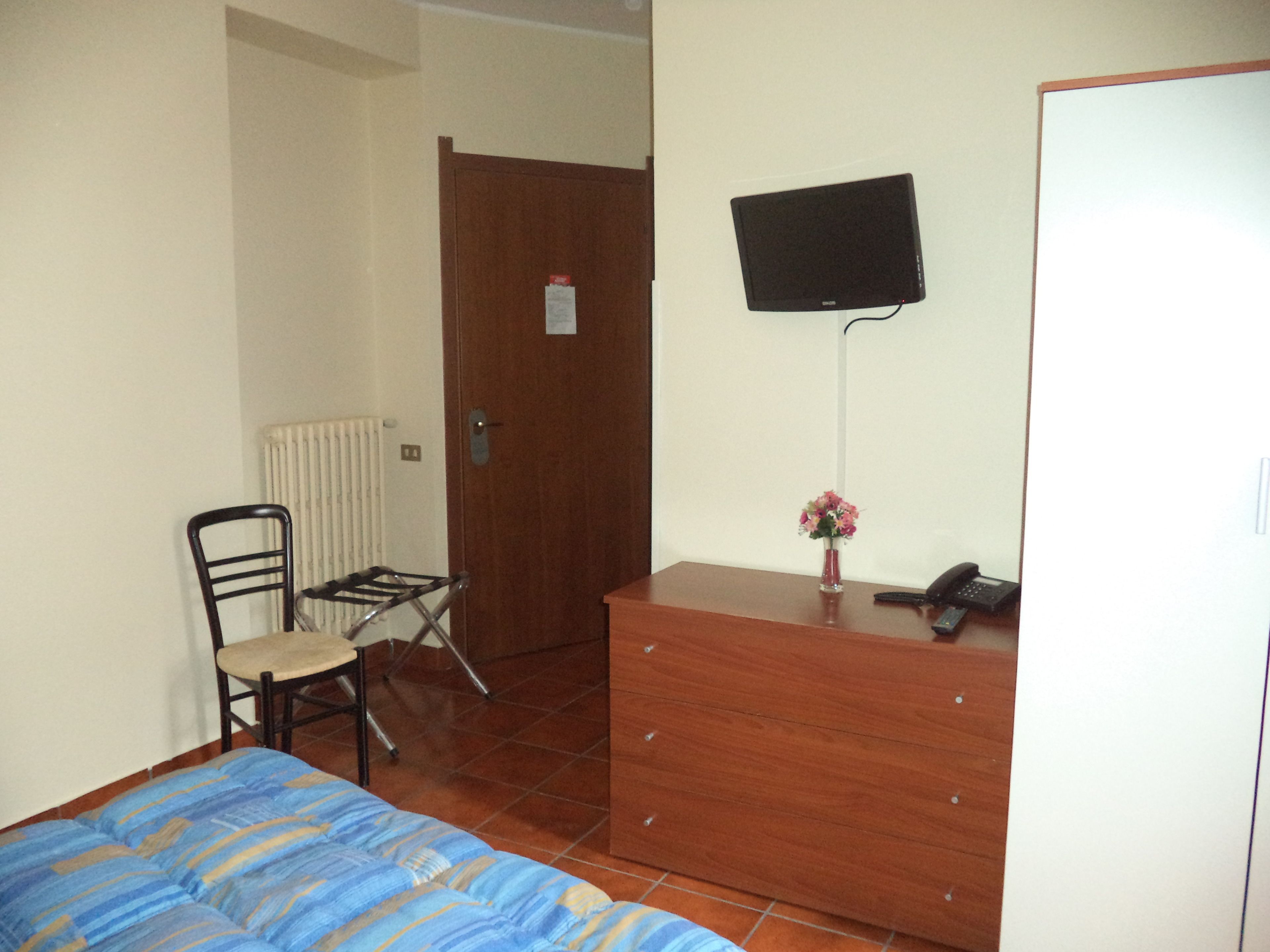 Una camera da letto con un letto, una cassettiera in legno, una sedia, un termosifone e un televisore fissato al muro.