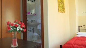 Un dormitorio con cama, una mesita de noche con un florero y una vista al baño a través de una puerta abierta, además de una obra de arte amarilla en la pared.