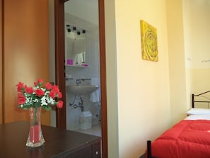 A bedroom with a bed, a nightstand with a vase of flowers, a bathroom visible through an open door, and a yellow artwork on the wall. - Hotel Il Nibbio (Magreglio)