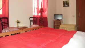 Un dormitorio con una cama grande, dos sillas, una mesita con un florero, un televisor sobre un mueble y una puerta.