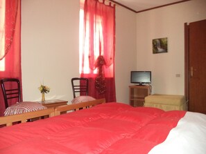 A bedroom with a large bed, two chairs, a small table with a vase, a television on a stand, and a door. - Hotel Il Nibbio (Magreglio)