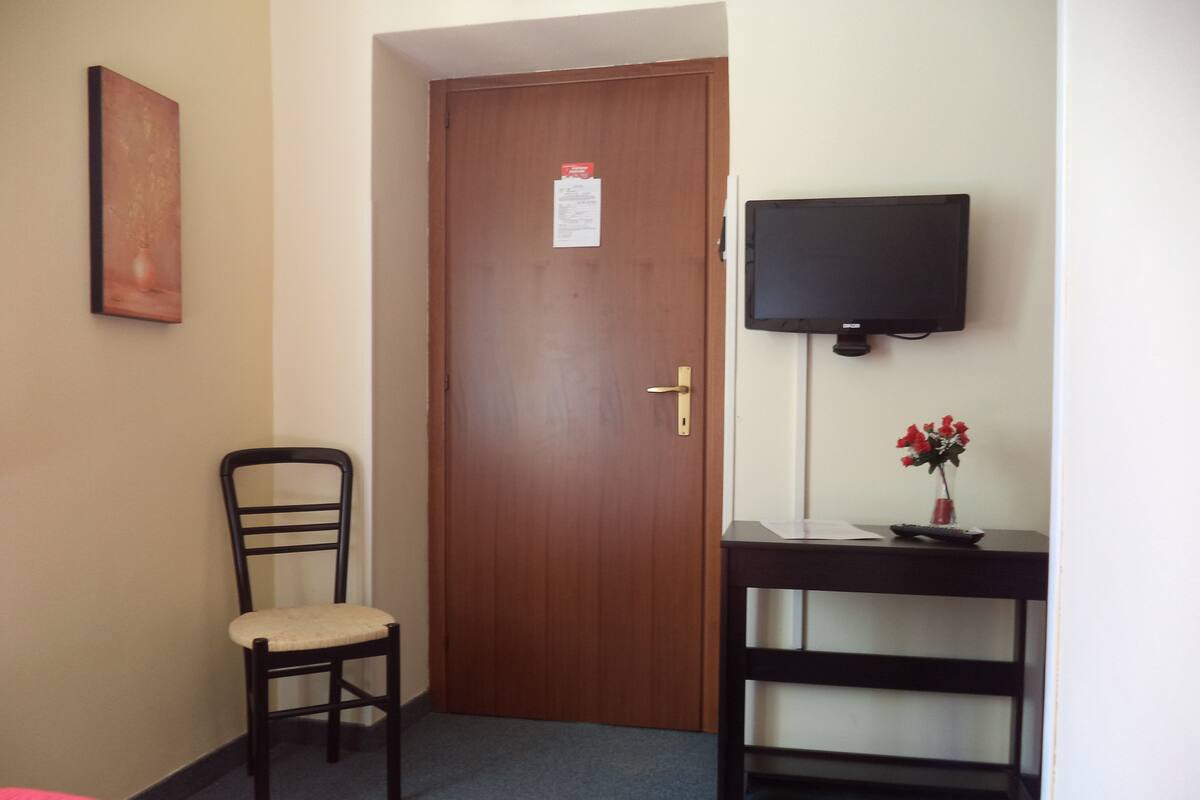 A hotel room with a wooden door, a chair, a small table with a vase of flowers, and a television on a stand.