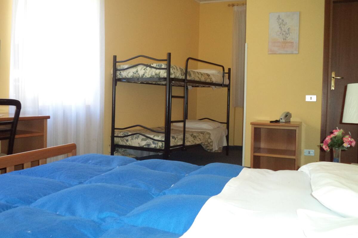 A room with bunk beds, a desk, a chair, a bedside table with a lamp, and a door.