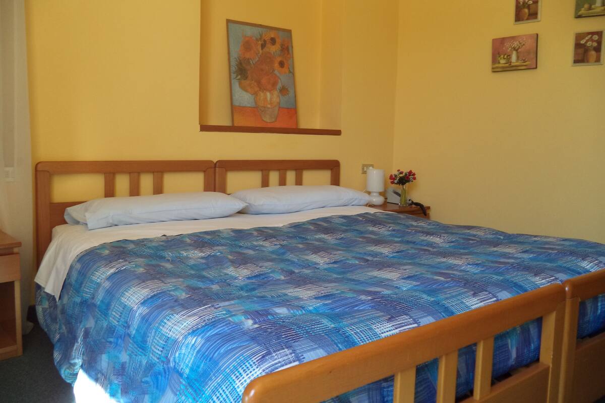 A bedroom with a wooden bed frame, a blue patterned bedspread, and a single white bedside table with a lamp and a vase of flowers.
