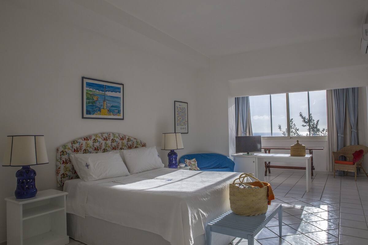 A hotel room with a bed, a desk, a chair, a window with curtains, and a painting on the wall.