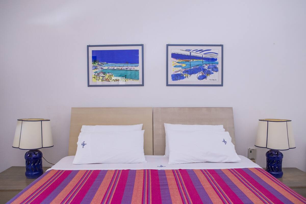 A hotel room with a bed, two white pillows, a striped blanket, two lamps, and two framed artworks on the wall.