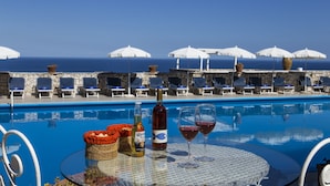 A poolside table with drinks, a bottle, and a basket, overlooking a pool with lounge chairs and umbrellas. - Hotel Piccolo Mondo (Castro)