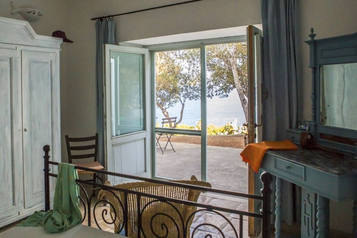 A room with a view of the sea, a dining area with a table and chairs, and a bed with a headboard.
