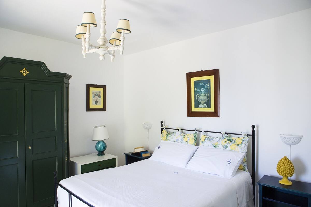 A neatly arranged bedroom with a bed, a green wardrobe, a chandelier, and two bedside tables with lamps and decorative items.