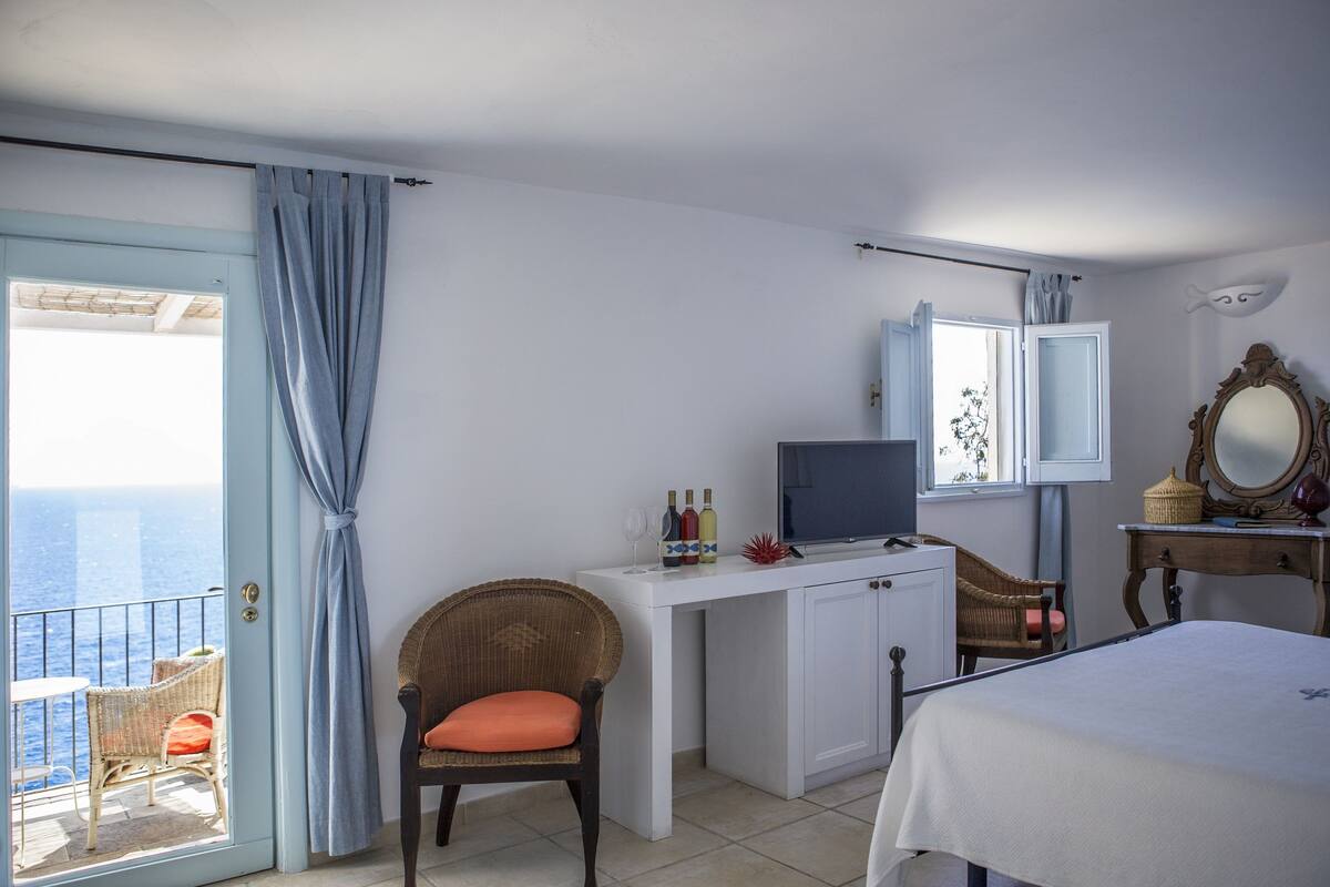 A bedroom with a bed, a desk with a television, a chair, a dresser, and a view of the sea through a balcony door.